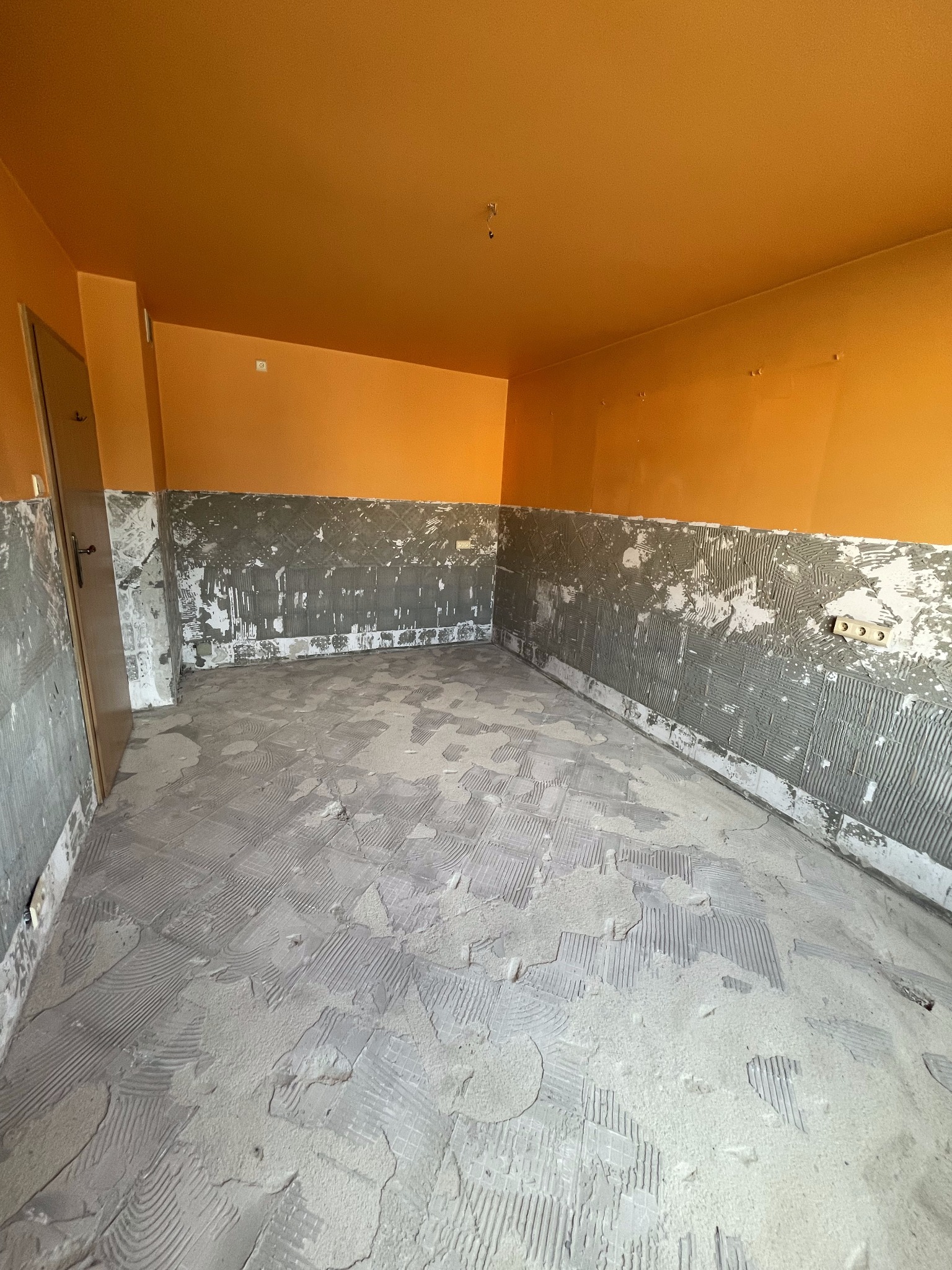 Pokój w trakcie prac przygotowawczych pod układanie płytek, widoczne ślady po kleju na ścianach i podłodze, pomarańczowy sufit.