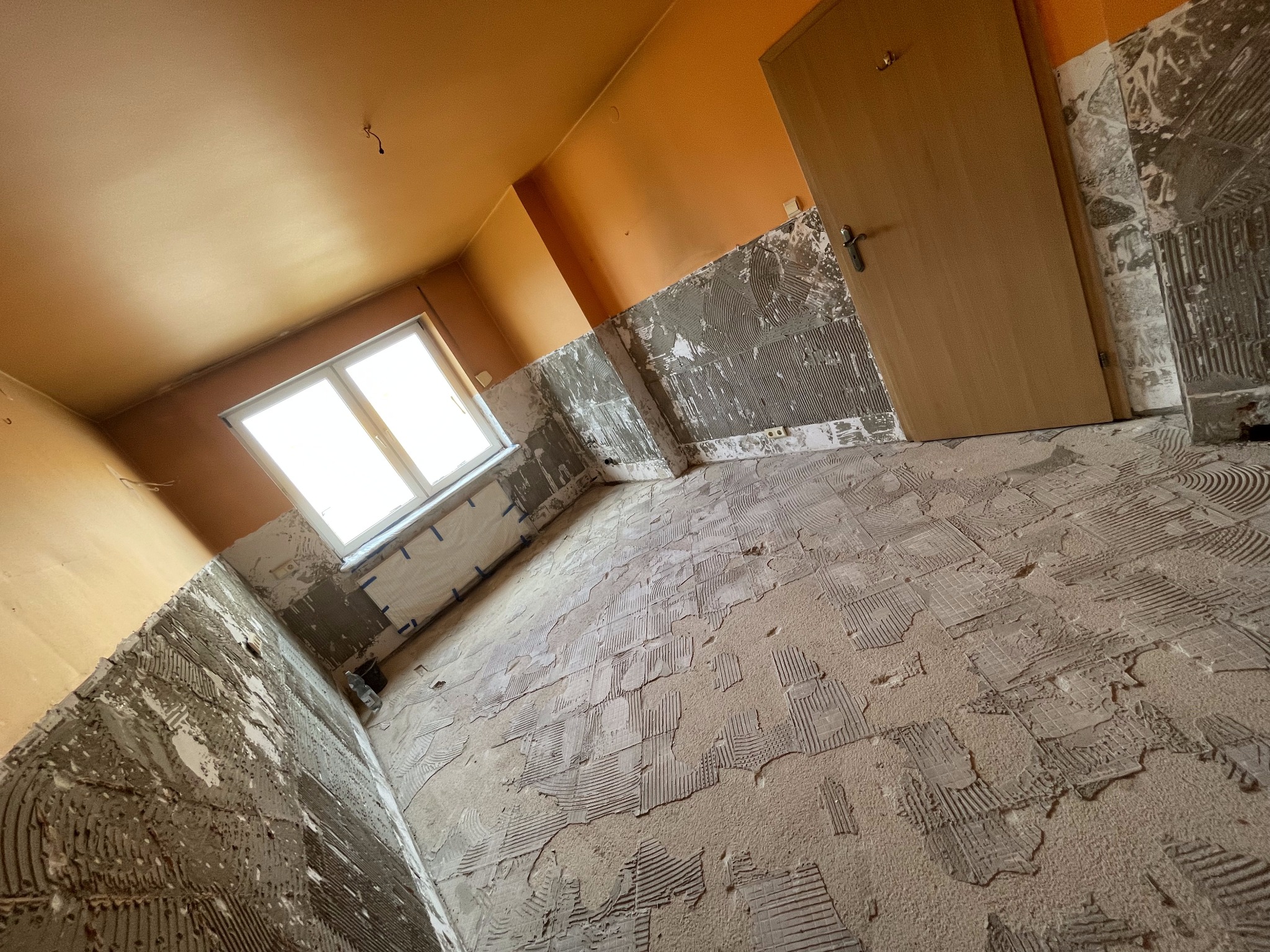 Pokój w trakcie przygotowania do położenia płytek, widoczne ślady kleju na ścianach i podłodze, pomarańczowe ściany, okno z zabezpieczeniem folią, drzwi drewniane.