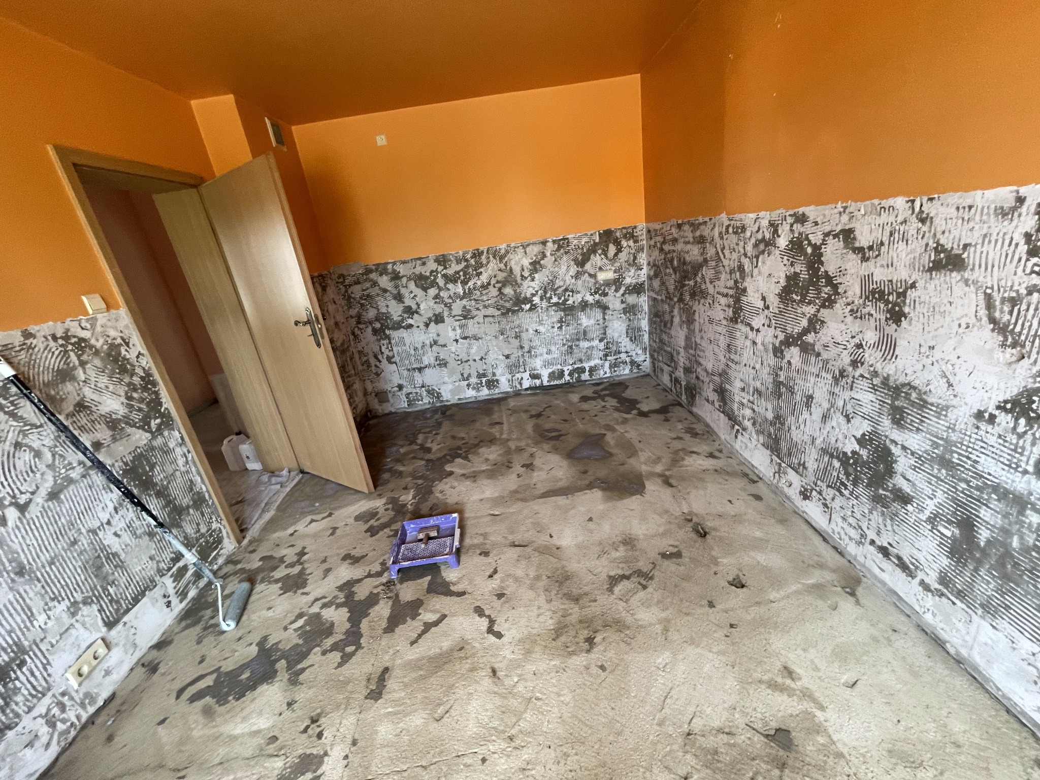 Pomarańczowe ściany w pokoju w trakcie przygotowywania podłoża, ściany częściowo pokryte warstwą wyrównującą, na podłodze ślady zaprawy, widoczny wałek i kuweta malarska.