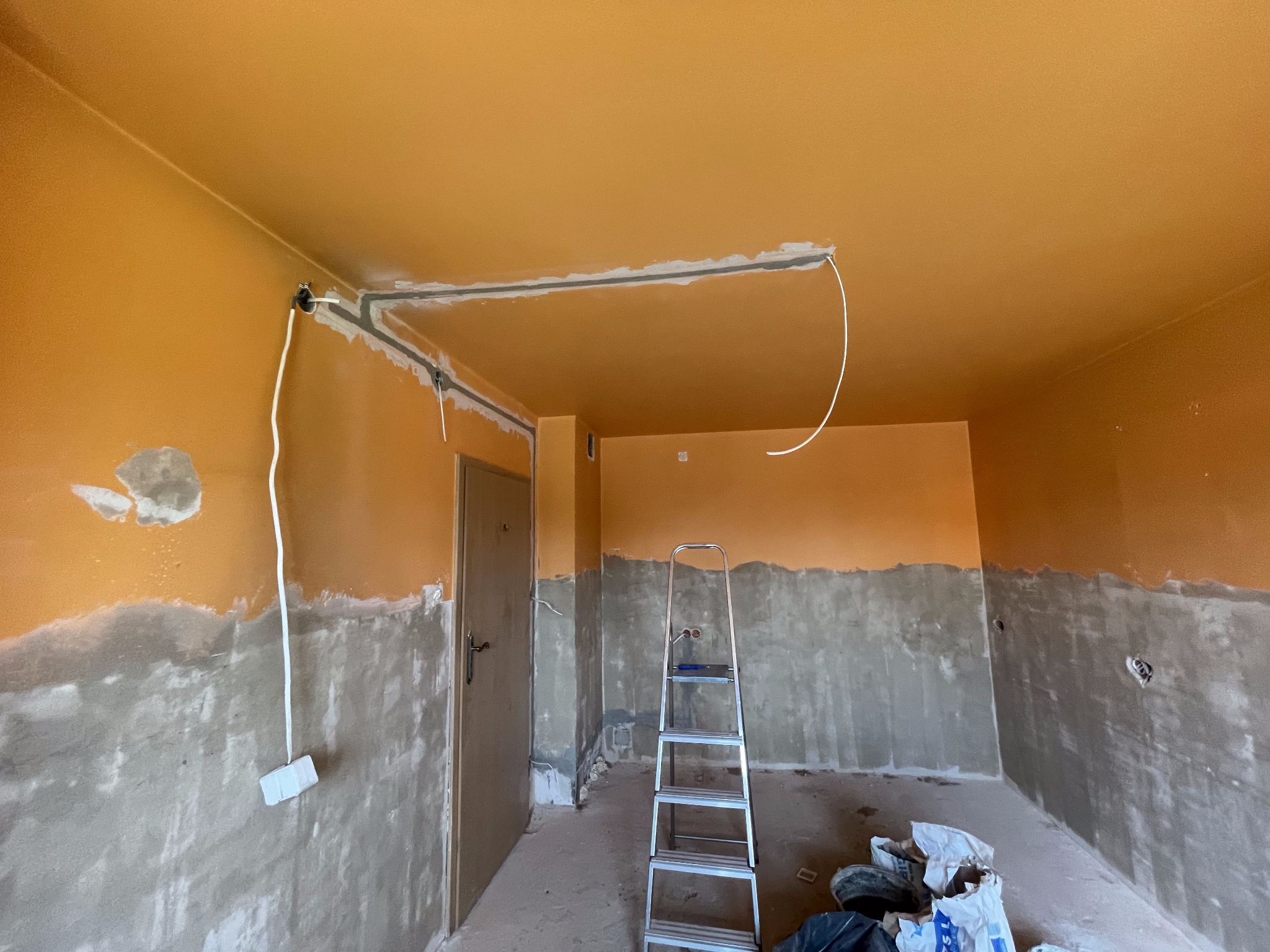 Pokój w trakcie prac przygotowawczych: pomarańczowa góra ścian i sufit, dolna część ścian w szarym tynku, widoczne przewody elektryczne, drabina aluminiowa i worki z materiałami budowlanymi...