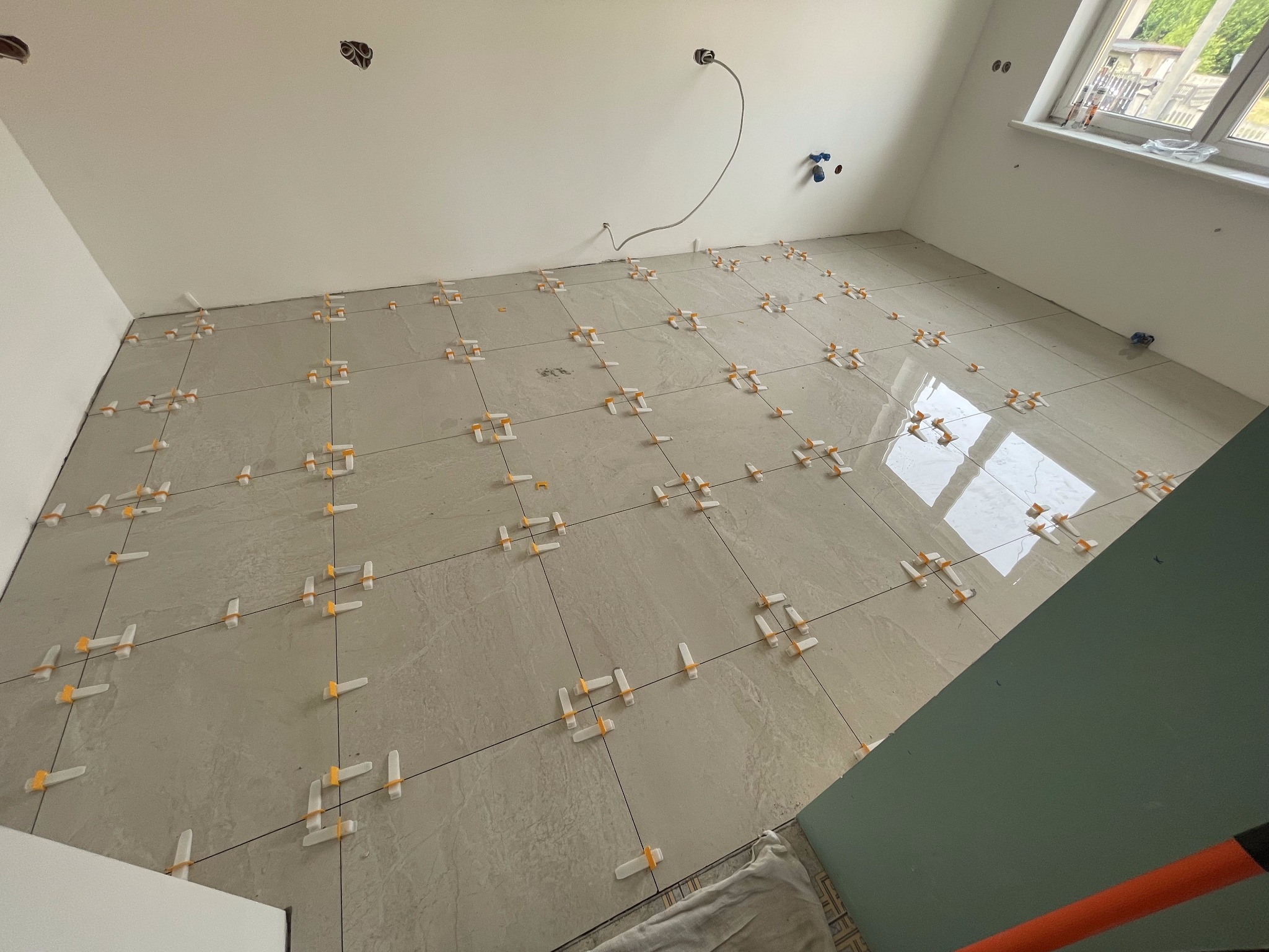 Jasnoszare płytki podłogowe w trakcie układania, z widocznymi krzyżykami dystansowymi, kabel wystający ze ściany, fragment okna z widokiem na zewnątrz.