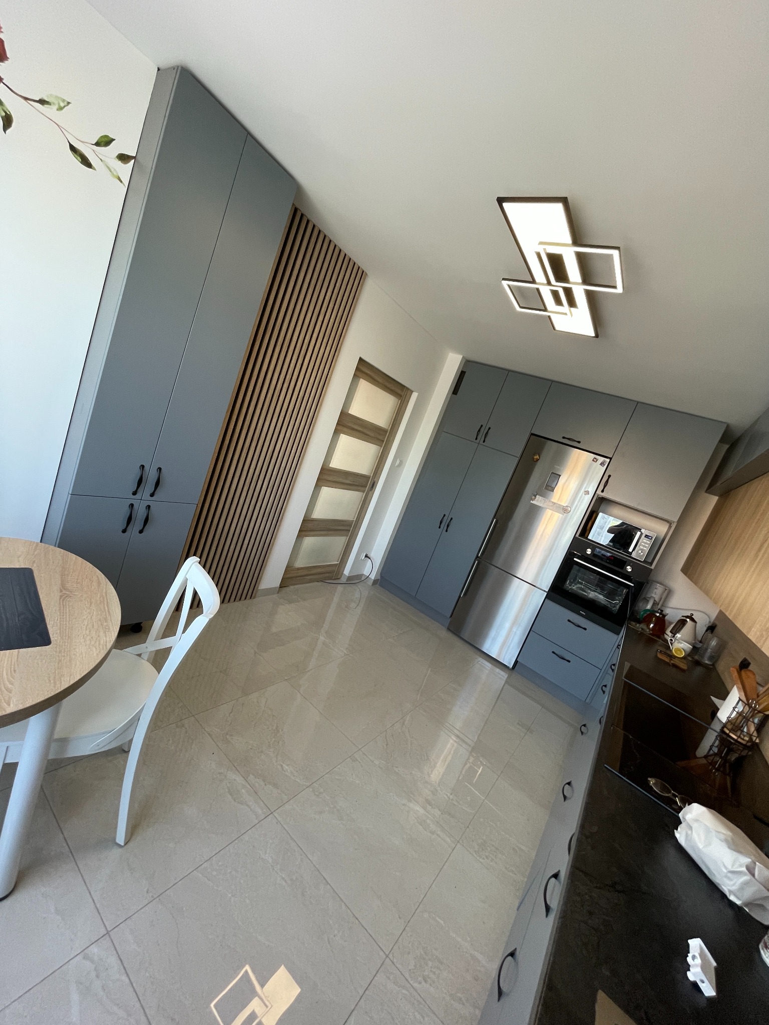 Nowoczesna kuchnia w odcieniach szarości z drewnianymi akcentami: szare szafki kuchenne, zabudowana lodówka, czarny blat, drewniana ściana z listew, okrągły stół z białym krzesłem, geometryczna...