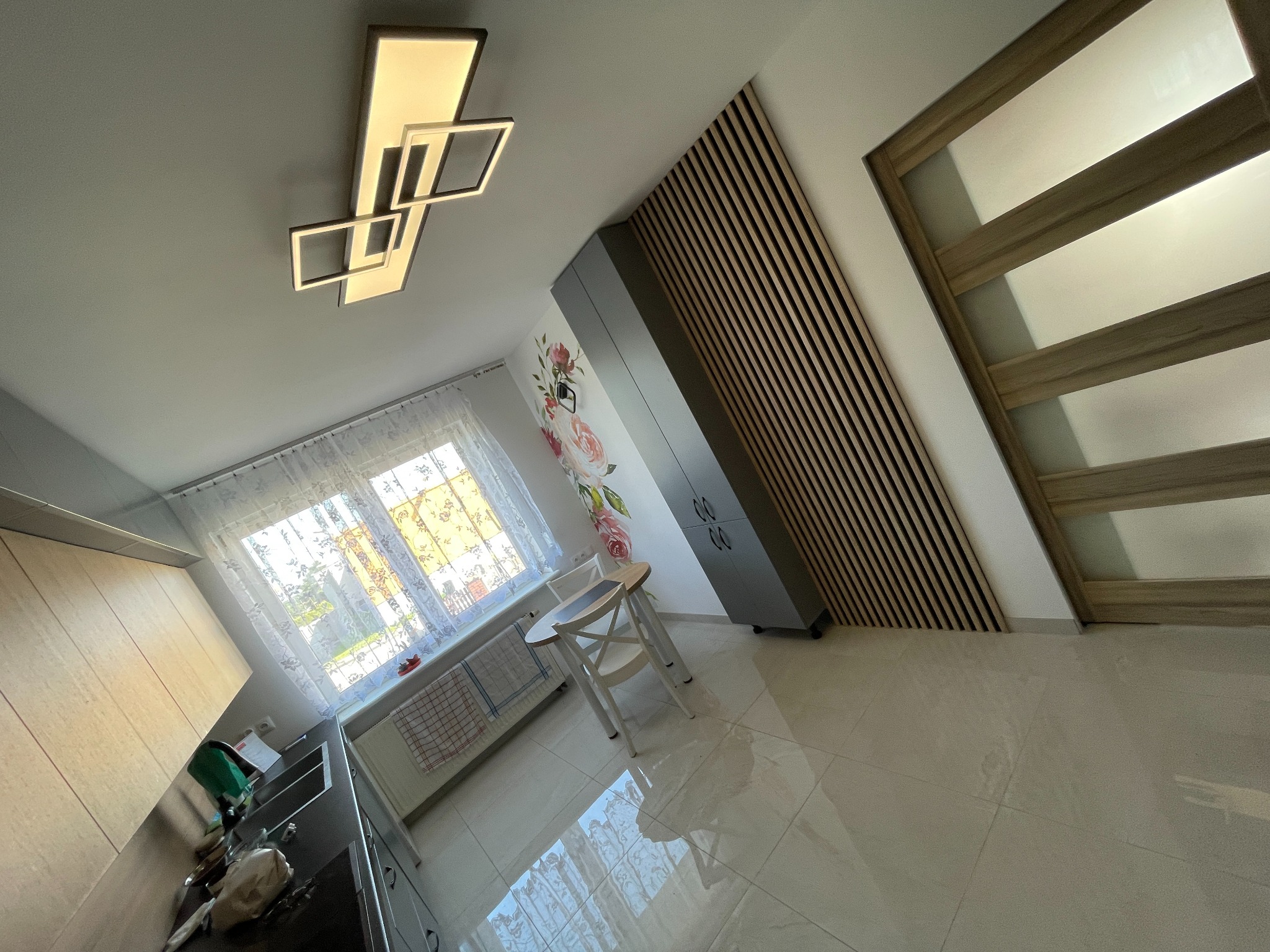 Nowoczesna kuchnia z drewnianą ścianą, wiszącą lampą w geometrycznym stylu i białymi płytkami podłogowymi. Widoczny fragment blatu kuchennego, okno z firanką oraz stół z krzesłami.