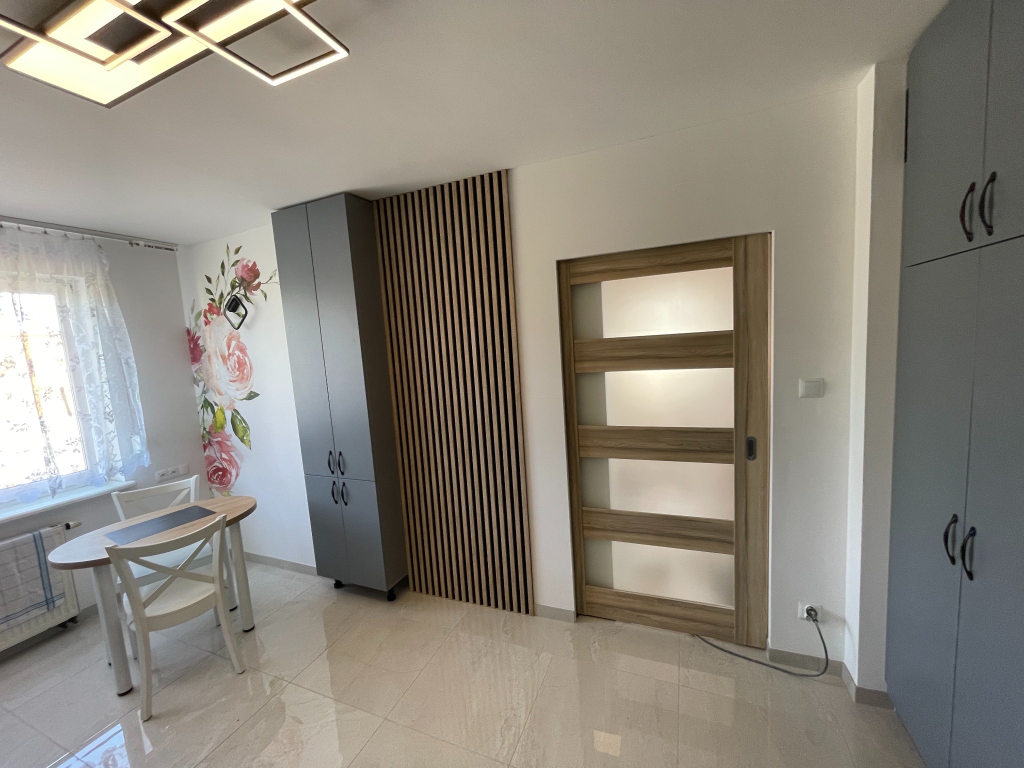 Jasne wnętrze po odświeżeniu: drewniane lamele na ścianie, szara szafa, drzwi z szybami i okleina w kolorze drewna, okrągły stół z krzesłami przy oknie z firanką i kwiatowy motyw naklejony na ścianie.