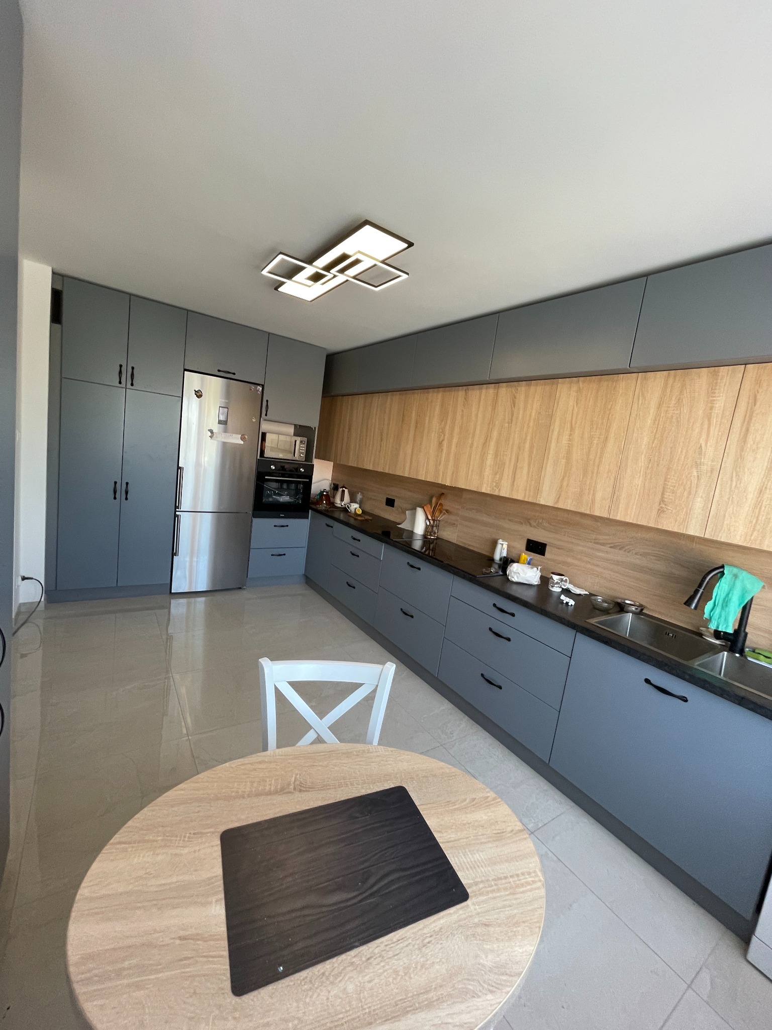 Nowoczesna kuchnia w odcieniach szarości i drewna z okrągłym stołem i białym krzesłem, oświetlona kwadratową lampą sufitową.