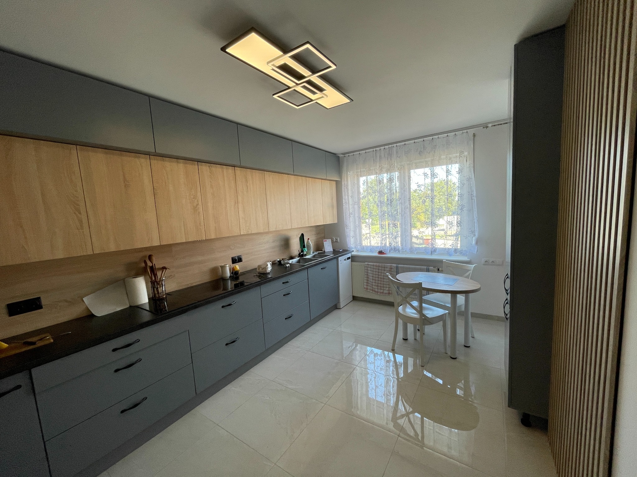 Nowoczesna kuchnia z szarymi szafkami dolnymi i drewnopodobnymi górnymi, czarnym blatem, białym stołem i krzesłami przy oknie, oraz dekoracyjną ścianą z drewnianych listewek.