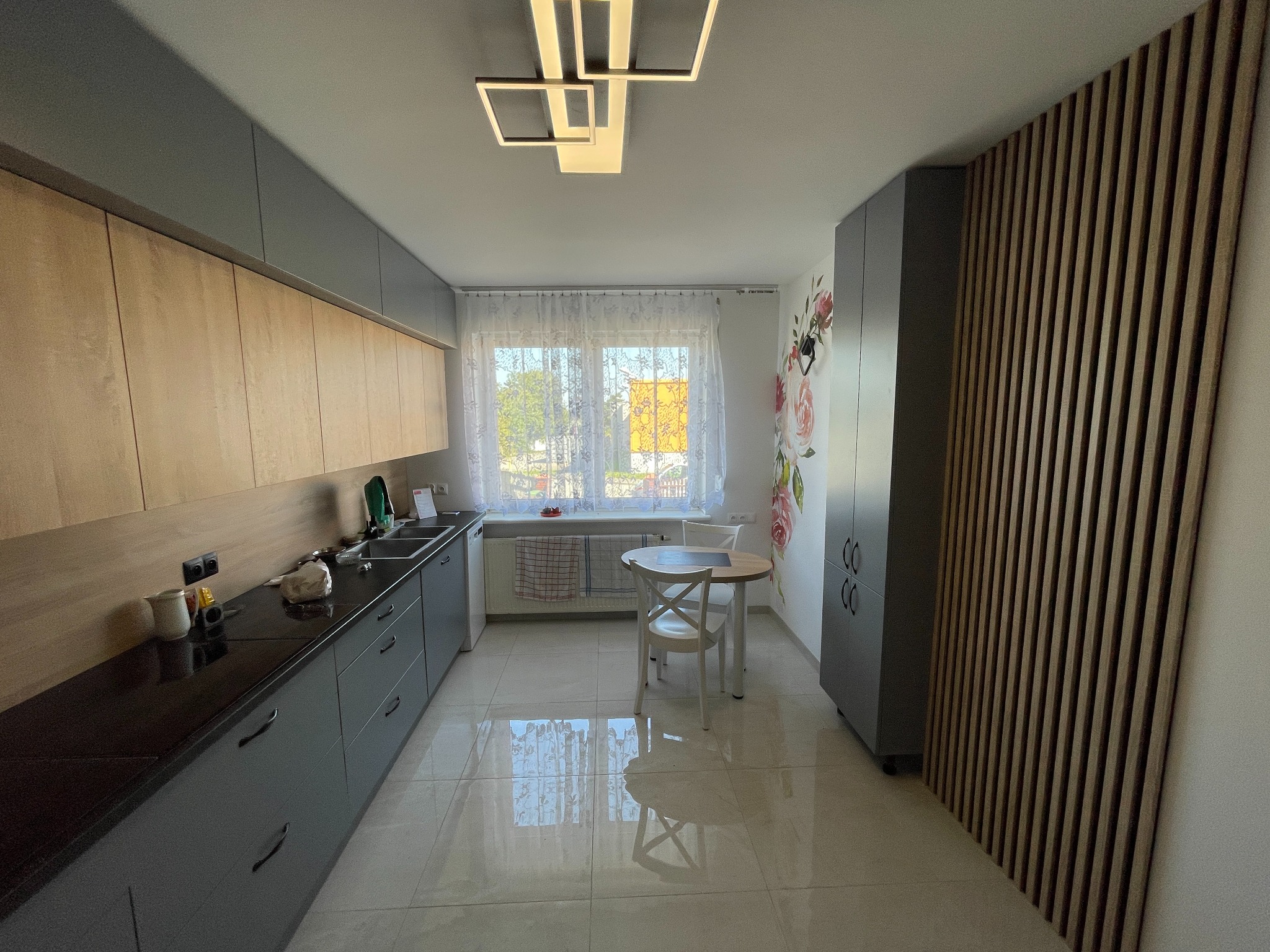 Nowoczesna kuchnia z szarymi szafkami, drewnianymi frontami, czarnym blatem, okrągłym stołem z białymi krzesłami, dekoracją kwiatową na ścianie i drewnianą ścianką działową z listew.