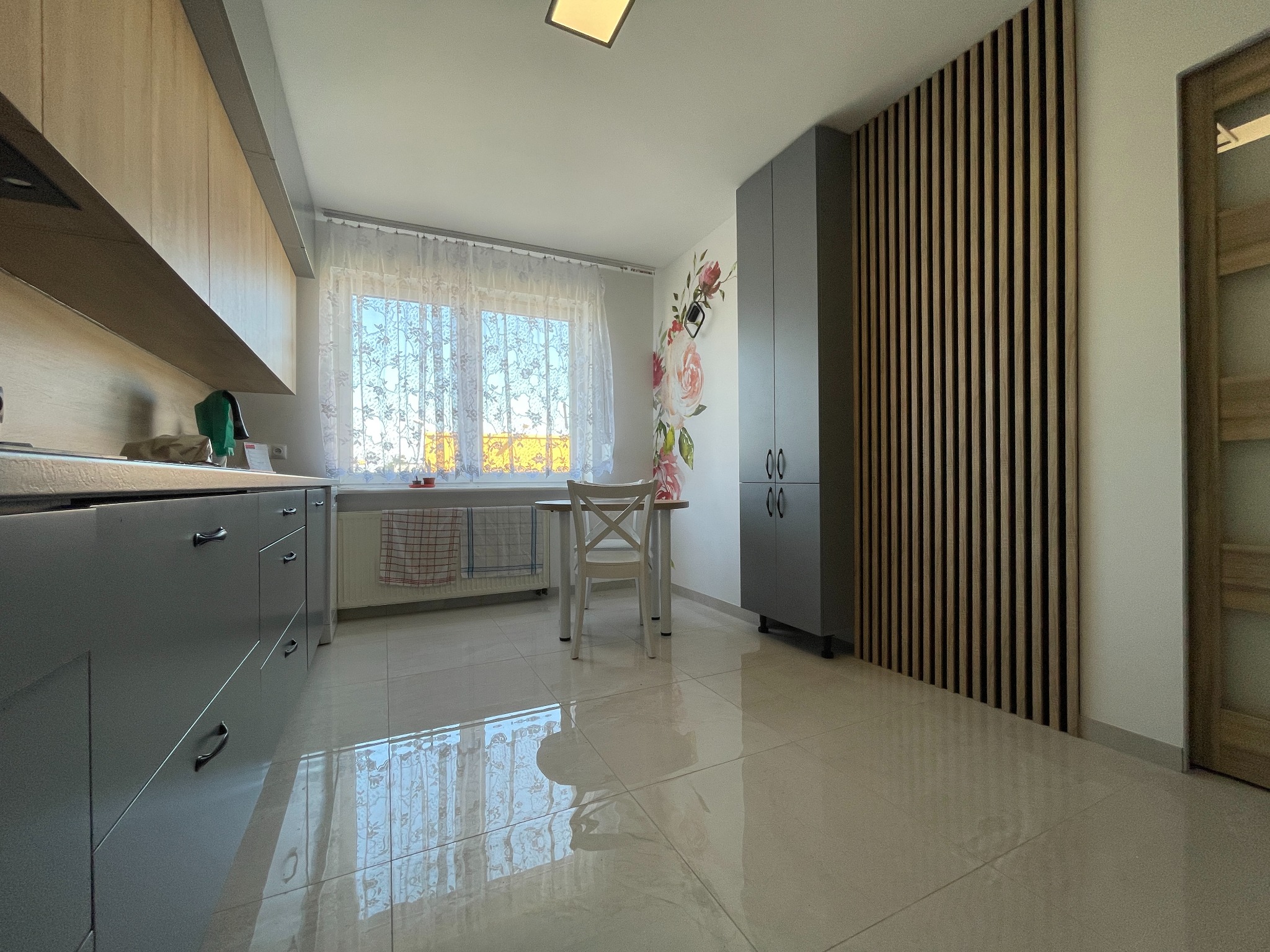 Jasna kuchnia z szarymi szafkami, drewnianymi lamelami na ścianie i białym stołem z krzesłem przy oknie z firankami.