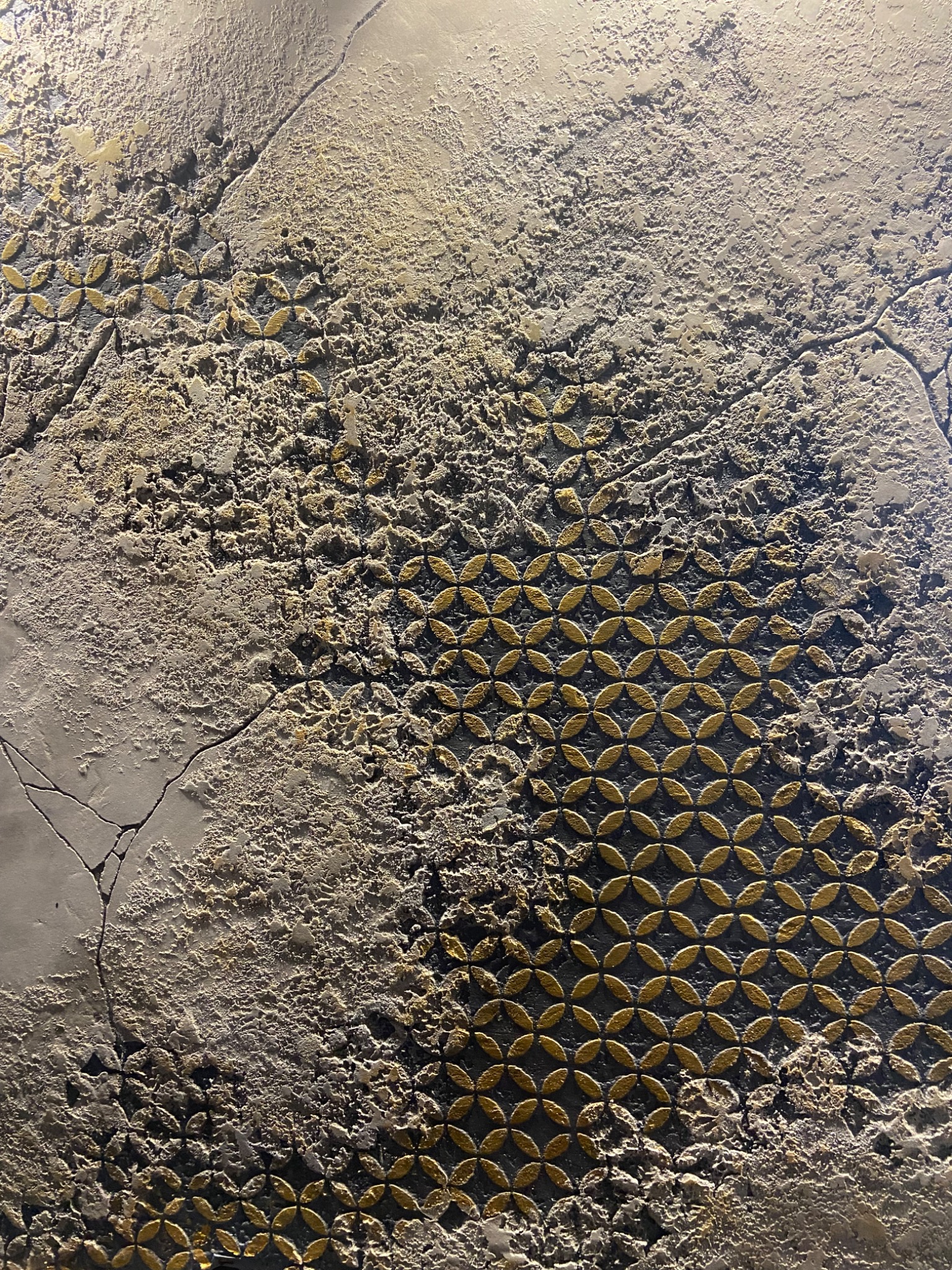 Dekoracyjna ściana z efektem betonu i złotymi, geometrycznymi wzorami, częściowo popękana, z widoczną fakturą i nierównościami powierzchni.