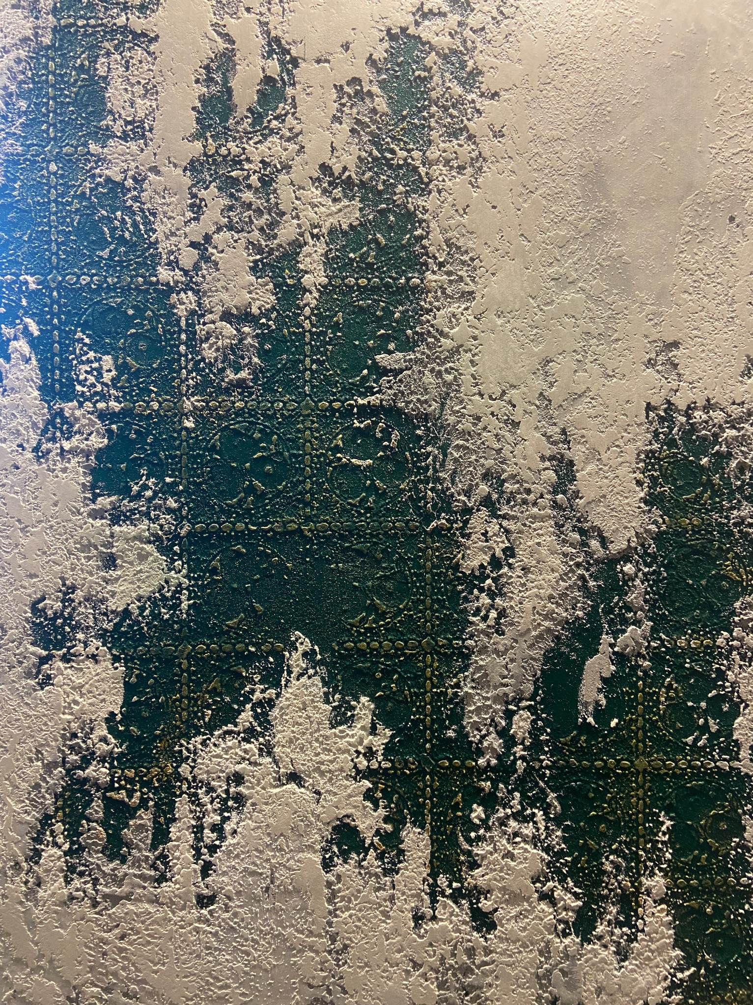 Nierównomiernie nałożony tynk dekoracyjny na ścianie, odsłaniający fragmenty ciemnozielonej, wzorzystej powierzchni pod spodem.