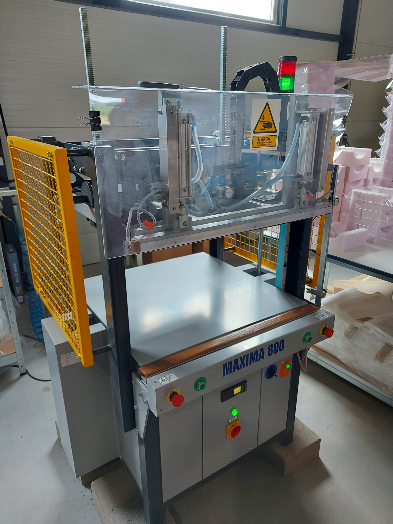 Maszyna przemysłowa Maxima 800 z żółtą siatką ochronną i przezroczystą osłoną, w tle zapakowane elementy z pianki.