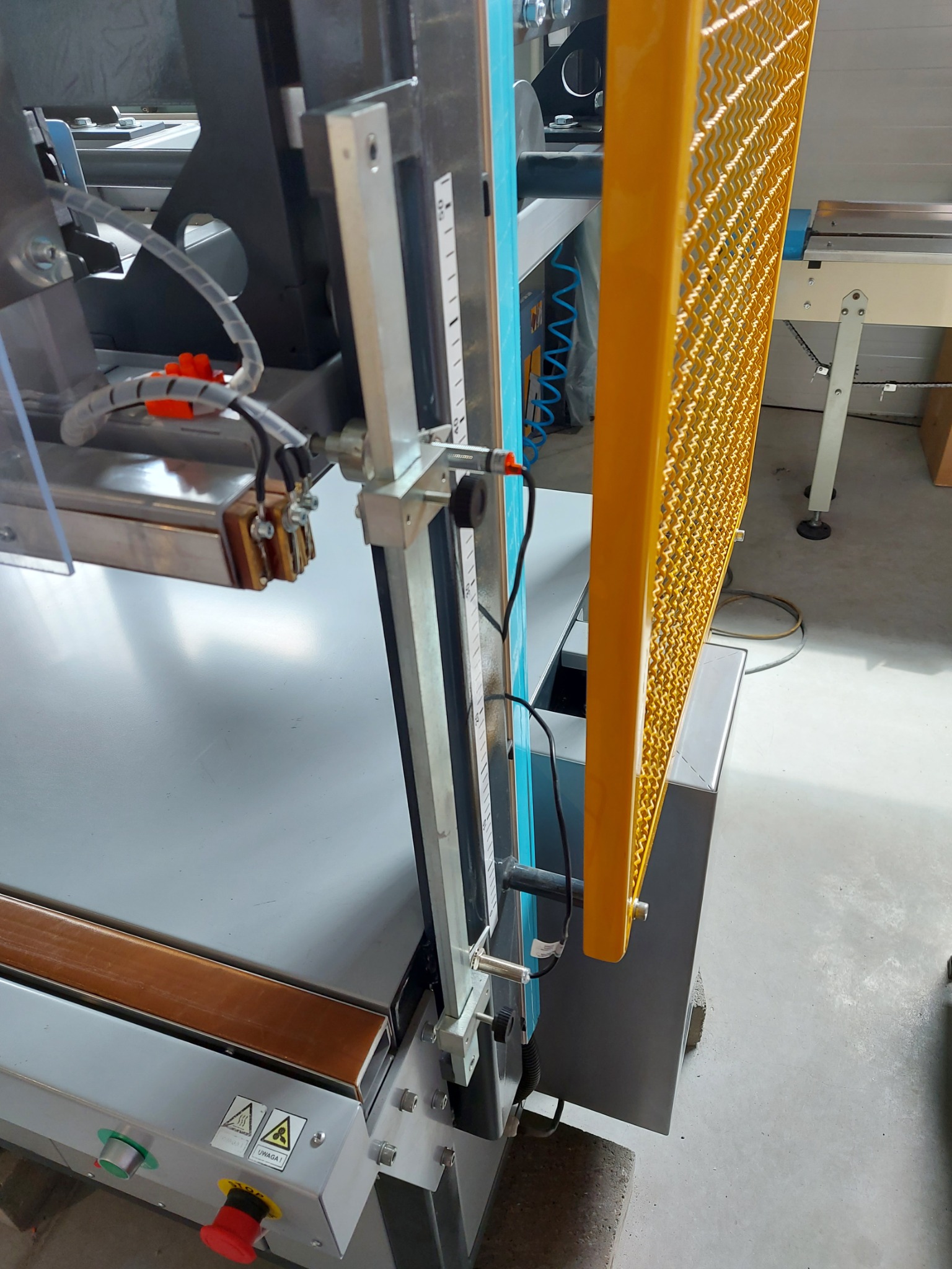 Fragment maszyny przemysłowej z metalową osłoną w kolorze żółtym, widoczna skala pomiarowa i elementy elektryczne.