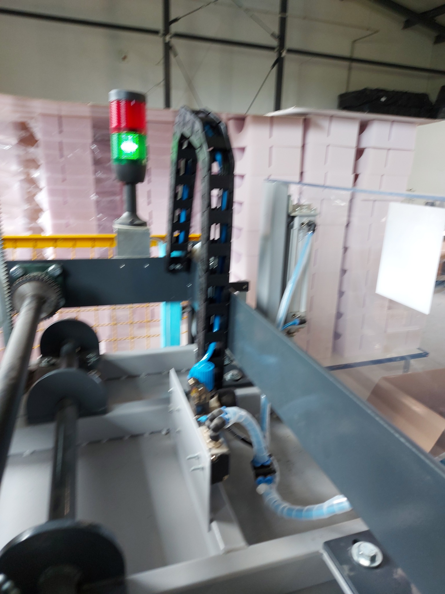 Fragment automatycznej linii produkcyjnej z widocznym sygnalizatorem świetlnym (czerwono-zielonym) i elementami pneumatyki, w tle stosy zapakowanych produktów.