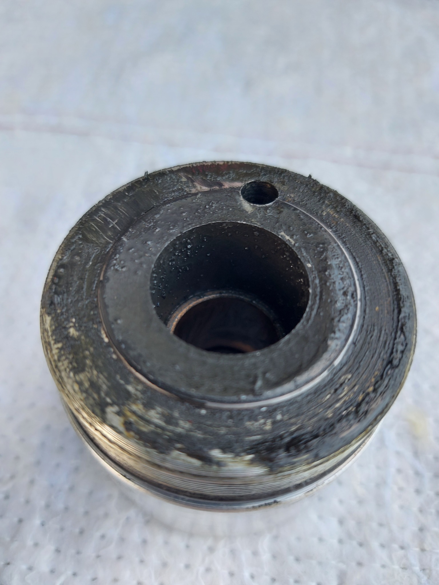 Zużyty, metalowy cylinder z otworem, pokryty smarem i brudem, leżący na białej powierzchni