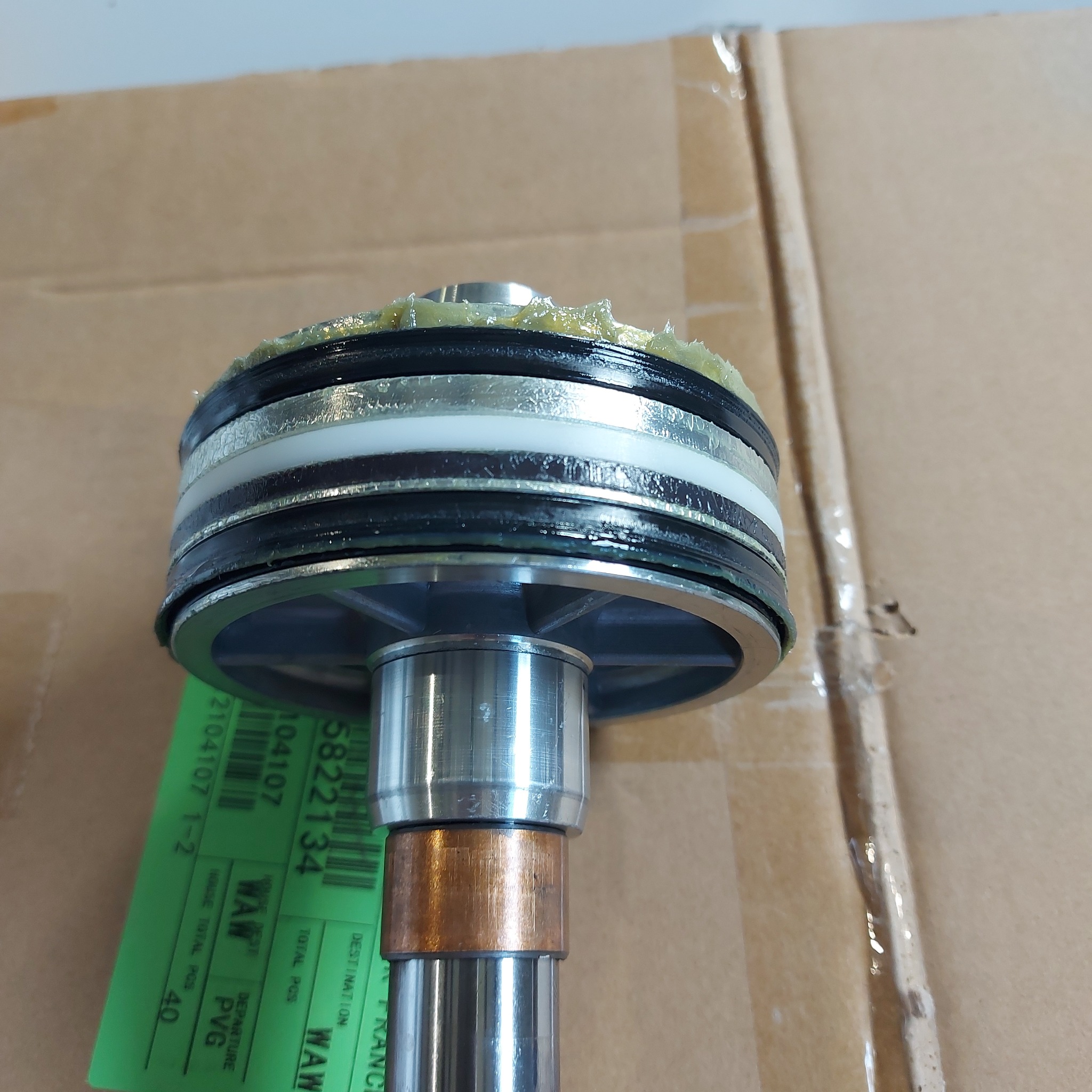 Część maszyny z elementami gumowymi, metalowymi i plastikowymi, nasmarowana smarem, na tle kartonowego pudła z zieloną etykietą z numerami.