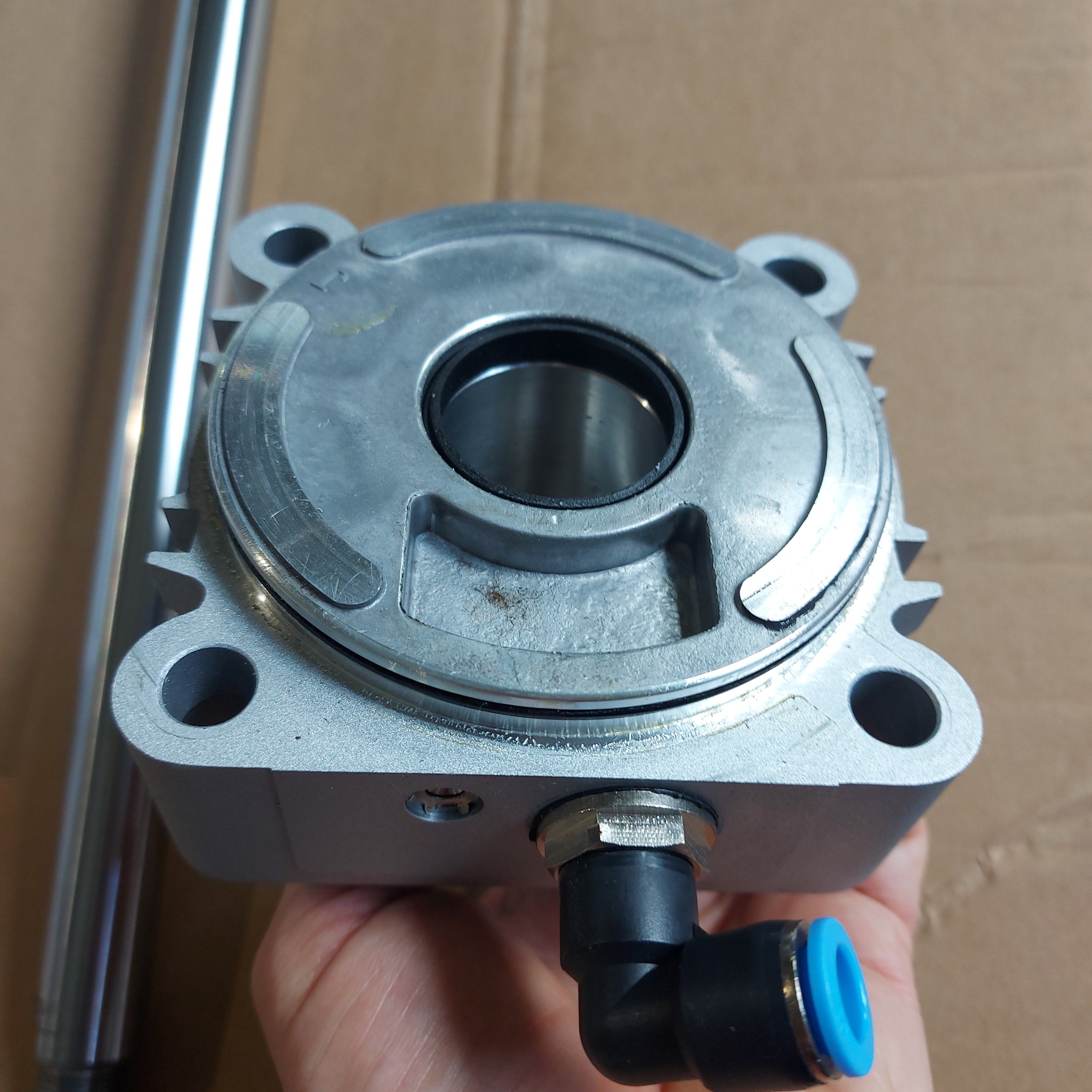 Aluminiowa głowica cylindra pneumatycznego z przyłączem, trzymana w dłoni na tle kartonu.