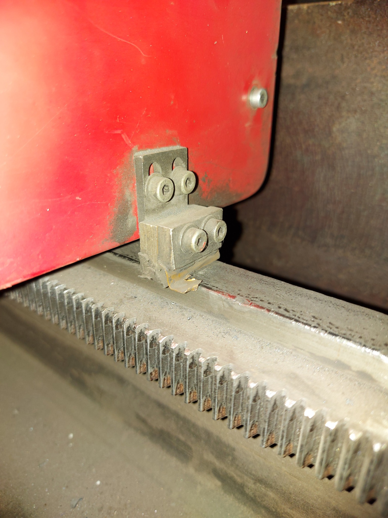 Metalowa listwa z zębami i przymocowany do niej element mocujący z czterema śrubami, część czerwonej maszyny w tle.