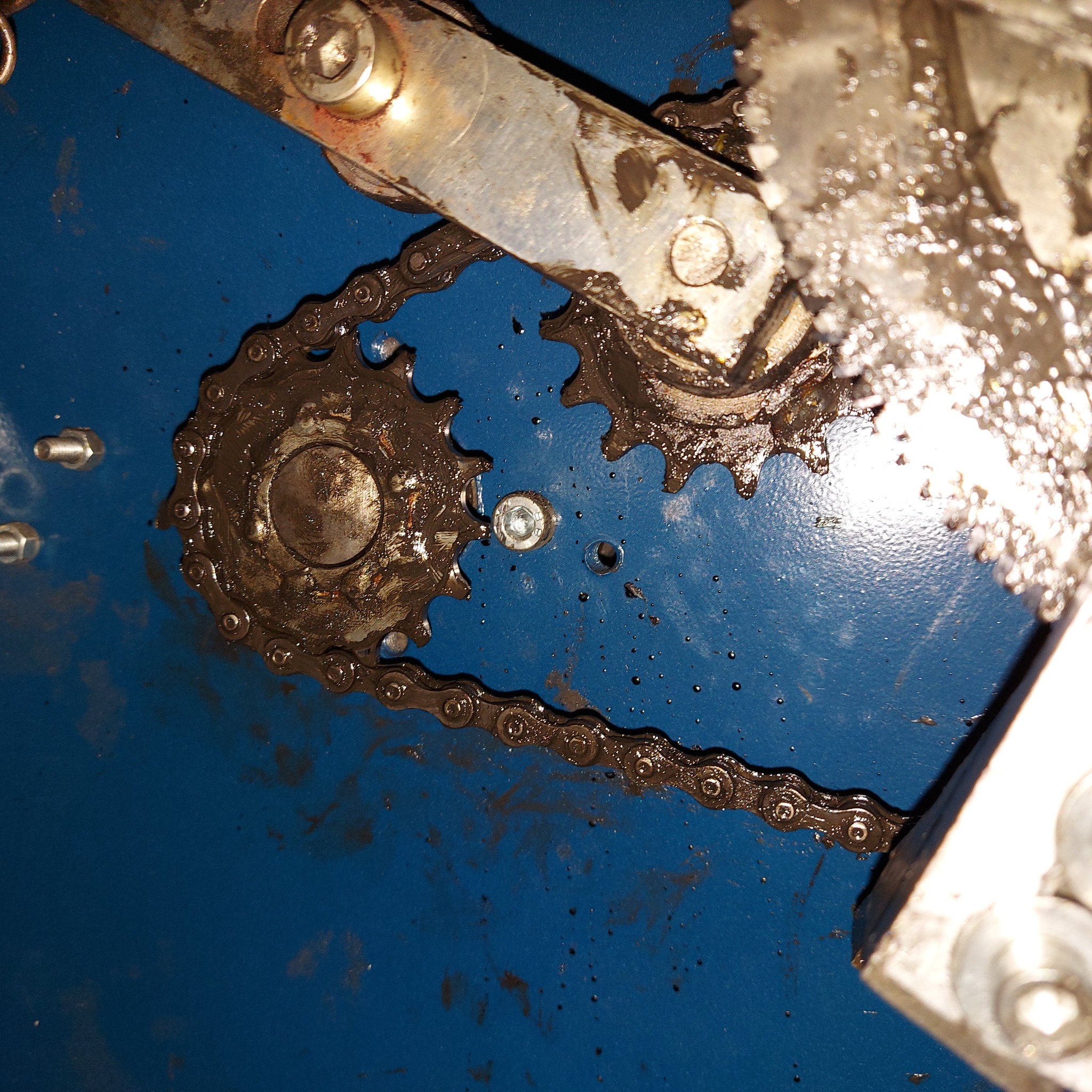 Brudny łańcuch i zębatki maszyny na niebieskim tle, widoczne ślady smaru i zabrudzeń, mocne oświetlenie z góry.