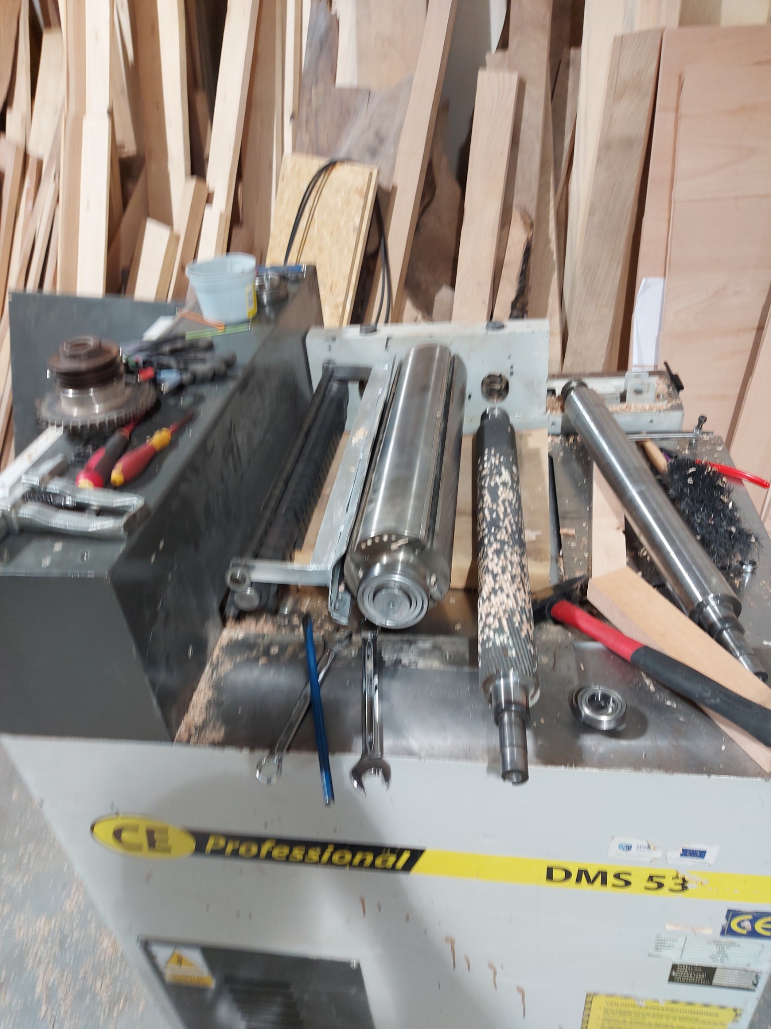 Rozłożona strugarka grubościówka DMS 53 z widocznymi wałami, narzędziami i drewnianymi wiórami na blacie roboczym, tło stanowią stosy drewnianych desek.