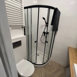 WOŹNIAK - Usługi Remontowe - Nowoczesna łazienka z kabiną prysznicową w czarnej ramie, wiszącą toaletą i grzejnikiem drabinkowym. Na podłodze płytki imitujące drewno ułożone w jodełkę.