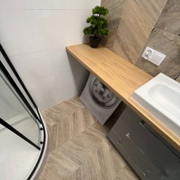 WOŹNIAK - Usługi Remontowe - Nowoczesna łazienka z kabiną prysznicową, umywalką wpuszczoną w blat z szafką, pralką ukrytą pod blatem i dekoracyjną rośliną w doniczce, wykończona płytkami imitującymi drewno w jodełkę.