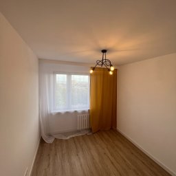 WOŹNIAK - Usługi Remontowe - Pusty pokój po remoncie z panelami podłogowymi w odcieniach brązu, pomalowanymi na biało ścianami, oknem z białymi firankami i żółtą zasłoną oraz nowoczesną czarną lampą sufitową z trzema żarówkami.