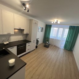 WOŹNIAK - Usługi Remontowe - Jasna kuchnia z czarnym blatem, białymi szafkami i drewnianą podłogą, widok na okno z zielonymi zasłonami, nowoczesne oświetlenie sufitowe i dekoracje.