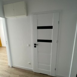 WOŹNIAK - Usługi Remontowe - Nowoczesne, białe drzwi wewnętrzne z czarnymi, poziomymi akcentami, zamontowane w jasnym pomieszczeniu z panelami podłogowymi i szafką na ścianie.