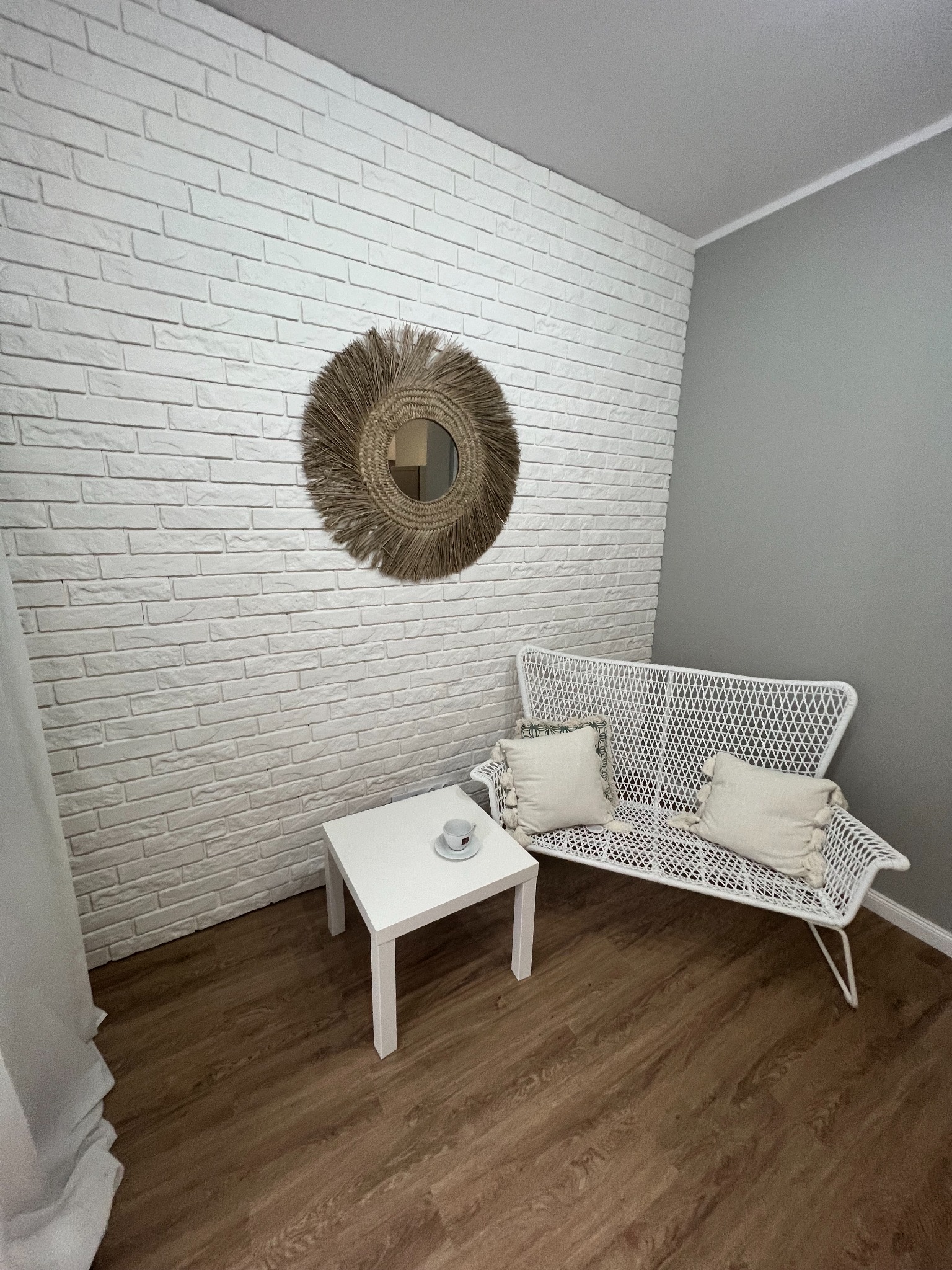 Jasne wnętrze z białą ceglaną ścianą, okrągłym lustrem w słomkowej ramie, białym stolikiem z filiżanką, wiklinową kanapą z poduszkami i drewnianą podłogą.
