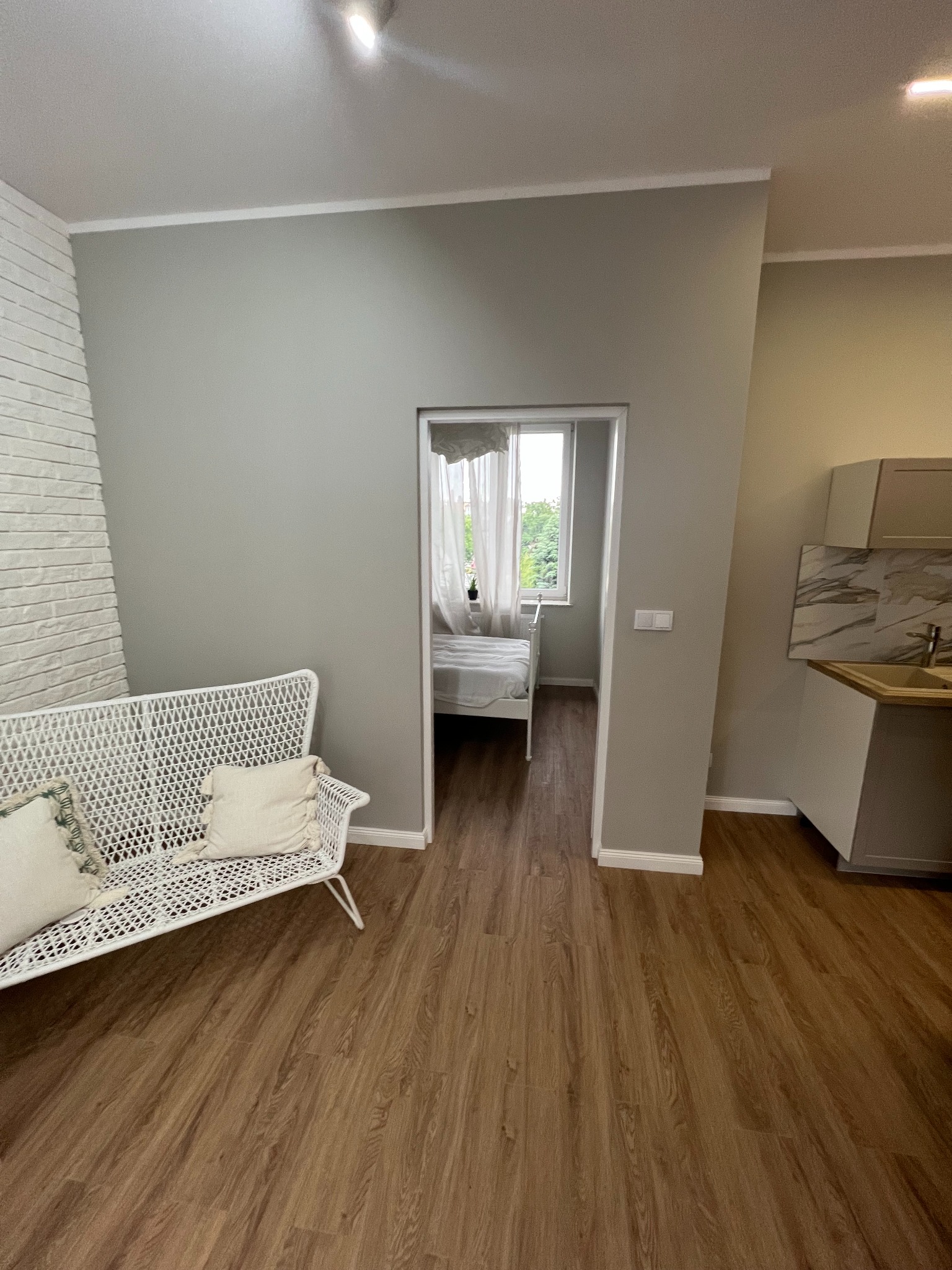 Jasne wnętrze mieszkania po remoncie: biała ceglana ściana, wiklinowa sofa z poduszkami, panele podłogowe w odcieniach brązu, widok na sypialnię przez otwarte drzwi, fragment aneksu kuchennego...