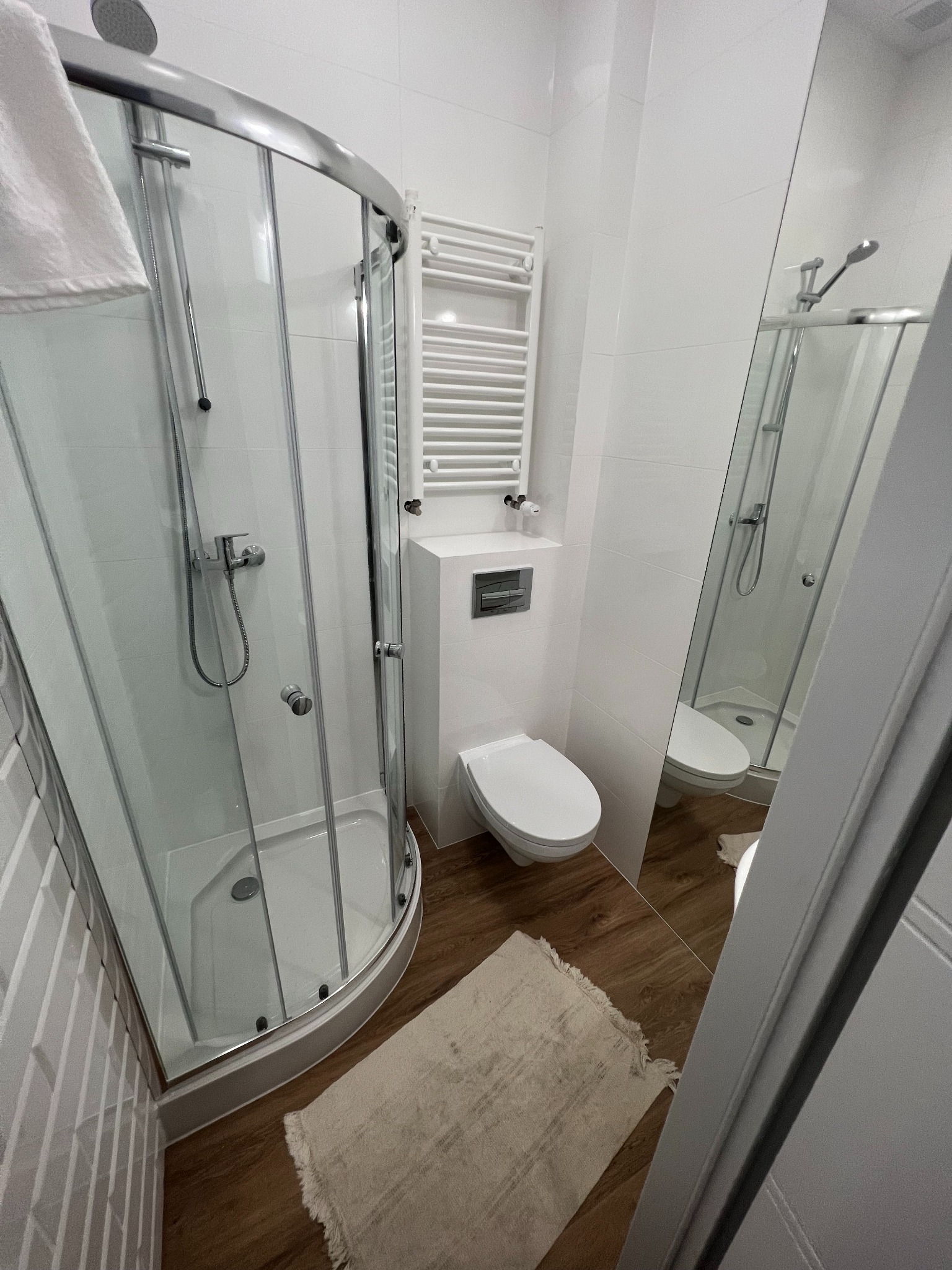 Nowoczesna łazienka z kabiną prysznicową z zaokrąglonymi drzwiami, toaletą podwieszaną, grzejnikiem drabinkowym i lustrem odbijającym kabinę, wykończona białymi płytkami i panelami podłogowymi...