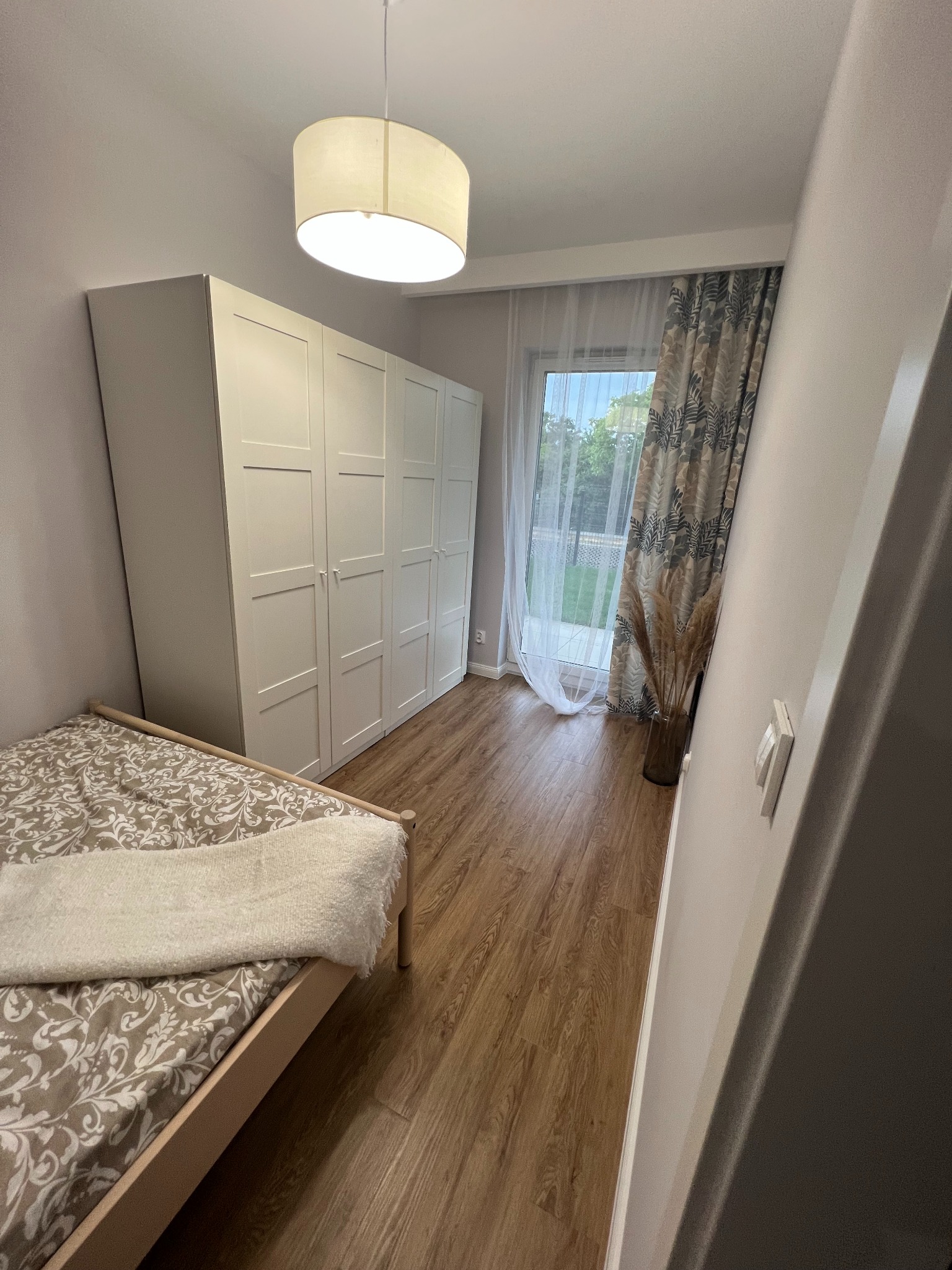 Jasne wnętrze sypialni z drewnianą podłogą, białymi szafami z czterema drzwiami, łóżkiem z wzorzystą pościelą i puszystym pledem, okno z firanką i zasłoną w neutralnych kolorach, oraz dekoracyjną...