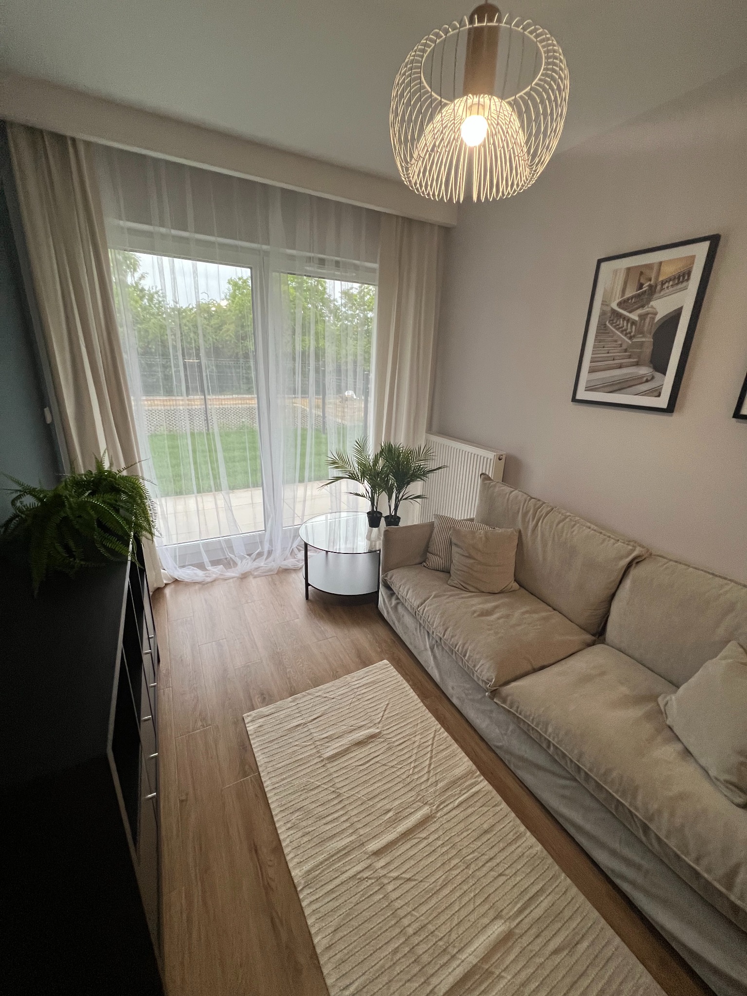 Jasny salon z drewnianą podłogą, beżową sofą, okrągłym stolikiem kawowym i dywanem w paski. Na ścianie wisi obraz w czarnej ramie przedstawiający schody. Lampa sufitowa z metalowych prętów...