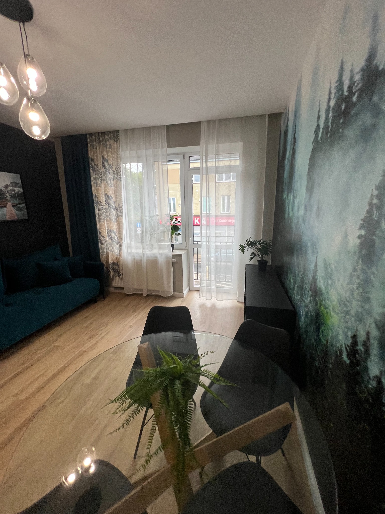 Nowoczesny salon z okrągłym szklanym stołem, czarnymi krzesłami, turkusową sofą i fototapetą z motywem leśnym na ścianie.