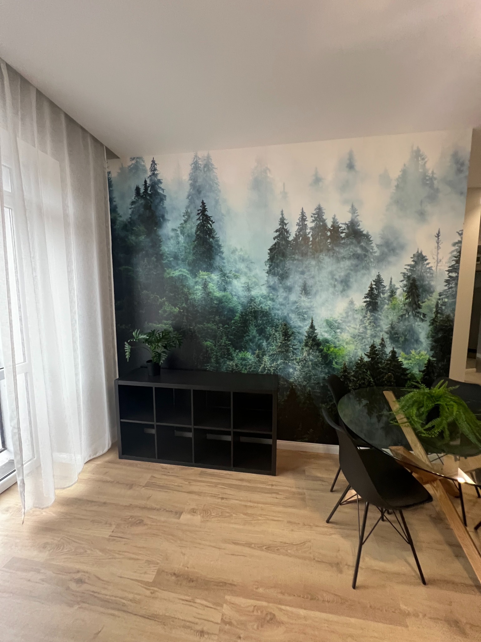 Salon z fototapetą przedstawiającą mglisty las, jasną podłogą z paneli, czarnym regałem i minimalistycznym stołem ze szklanym blatem.