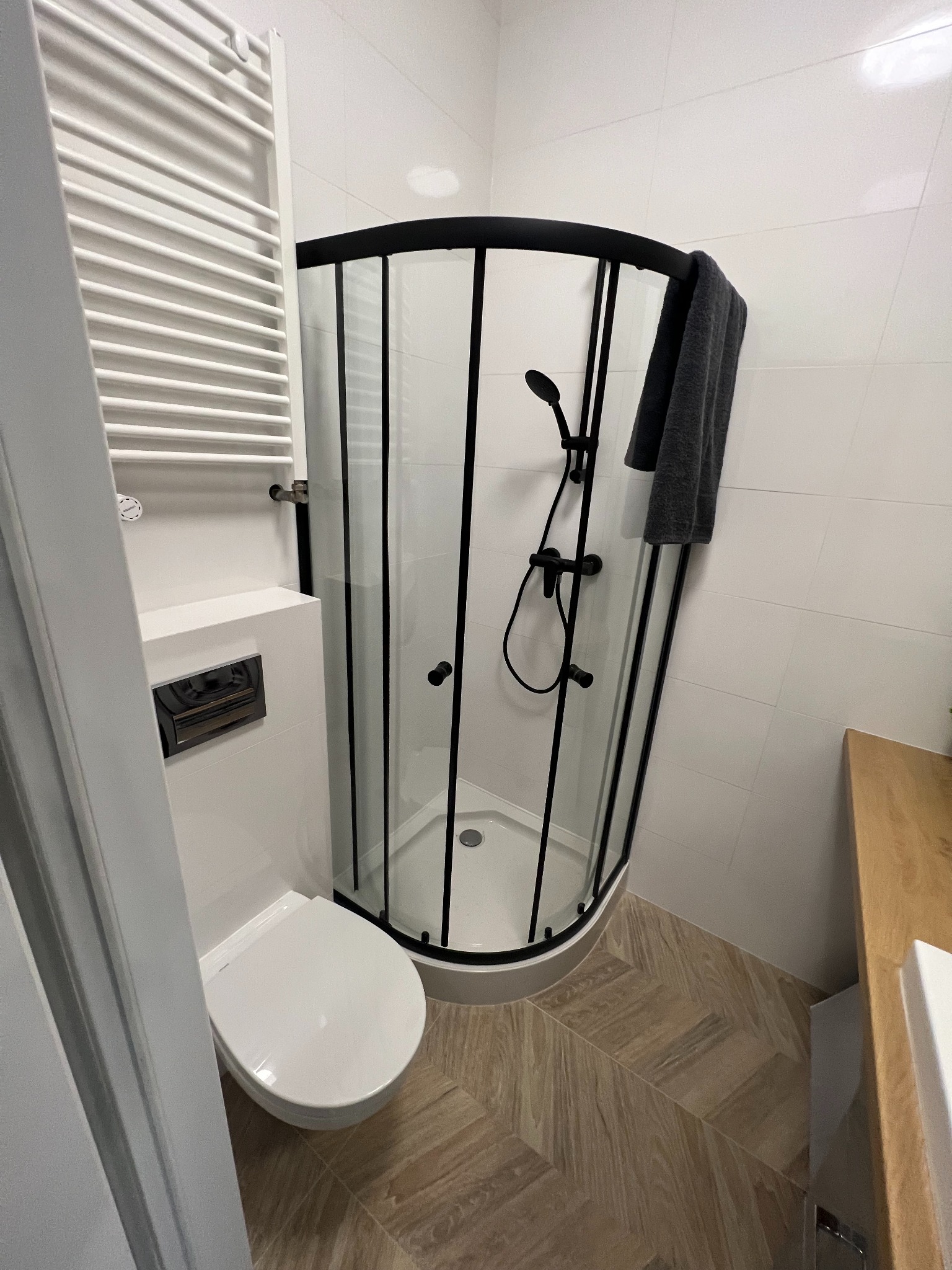 Nowoczesna łazienka z kabiną prysznicową w czarnej ramie, wiszącą toaletą i grzejnikiem drabinkowym. Na podłodze płytki imitujące drewno ułożone w jodełkę.