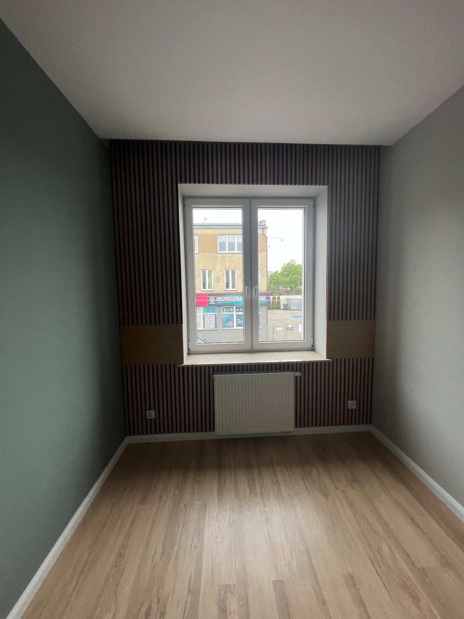 Pusty pokój po remoncie z panelami na podłodze, zieloną ścianą i dekoracyjnymi drewnianymi lamelami za oknem z grzejnikiem.