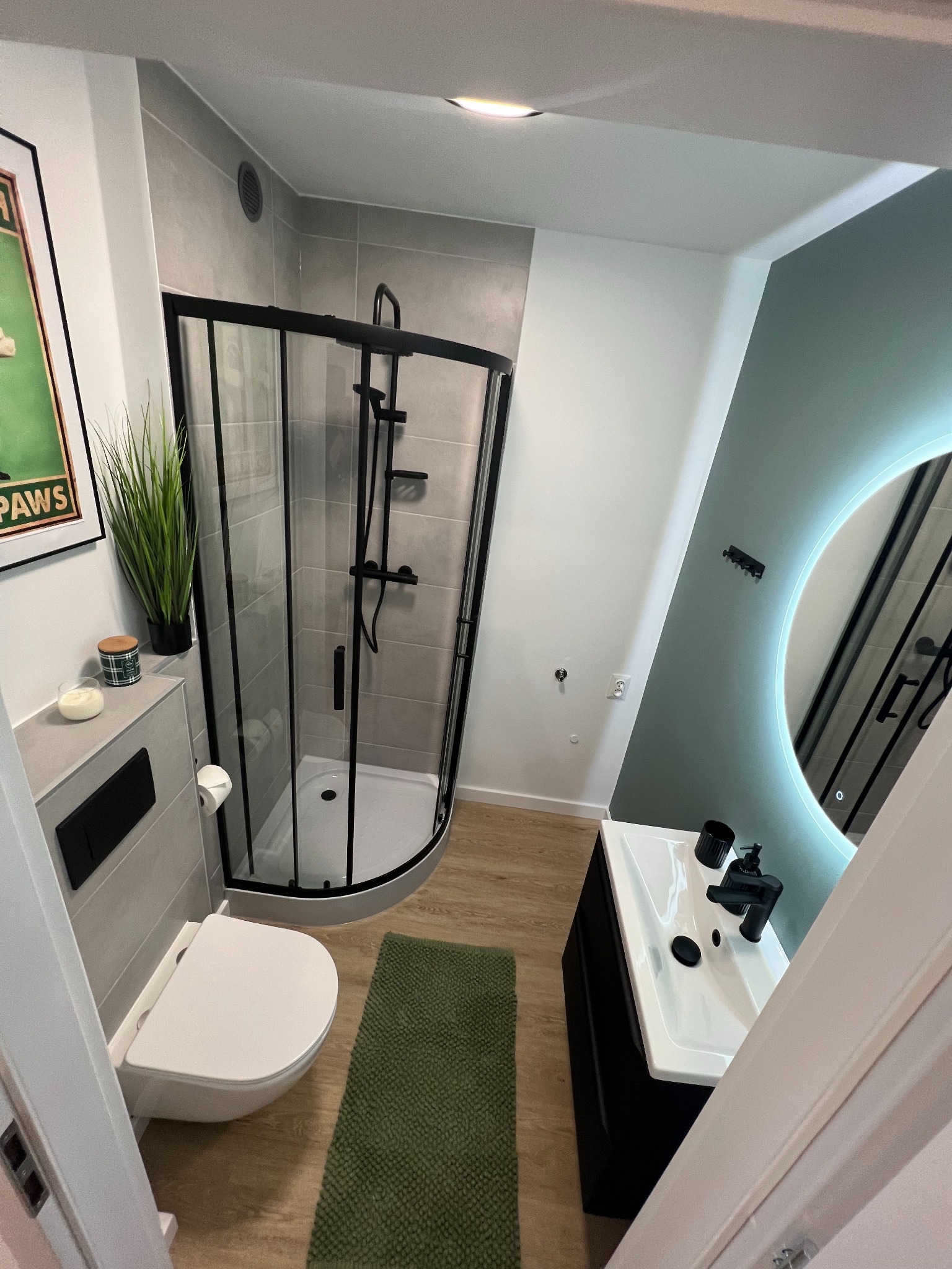 Nowoczesna łazienka z kabiną prysznicową typu walk-in z czarnymi profilami, umywalką z czarną armaturą, okrągłym lustrem z podświetleniem LED i zielonym akcentem kolorystycznym na ścianie.