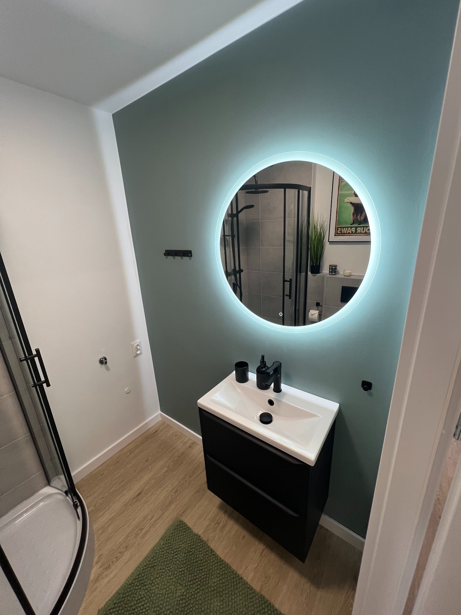 Nowoczesna łazienka z okrągłym lustrem podświetlanym LED, czarną armaturą i kabiną prysznicową z czarnymi profilami, zielona ściana i dywanik.