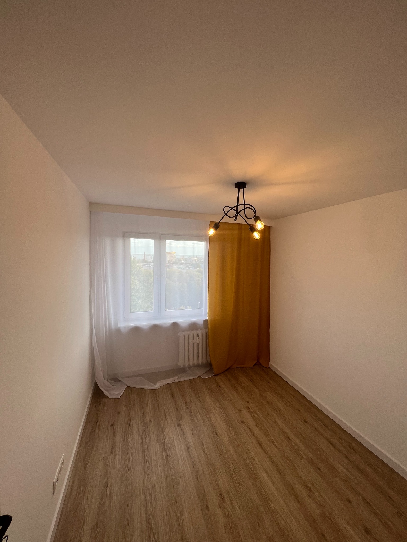 Pusty pokój po remoncie z panelami podłogowymi w odcieniach brązu, pomalowanymi na biało ścianami, oknem z białymi firankami i żółtą zasłoną oraz nowoczesną czarną lampą sufitową z trzema żarówkami.