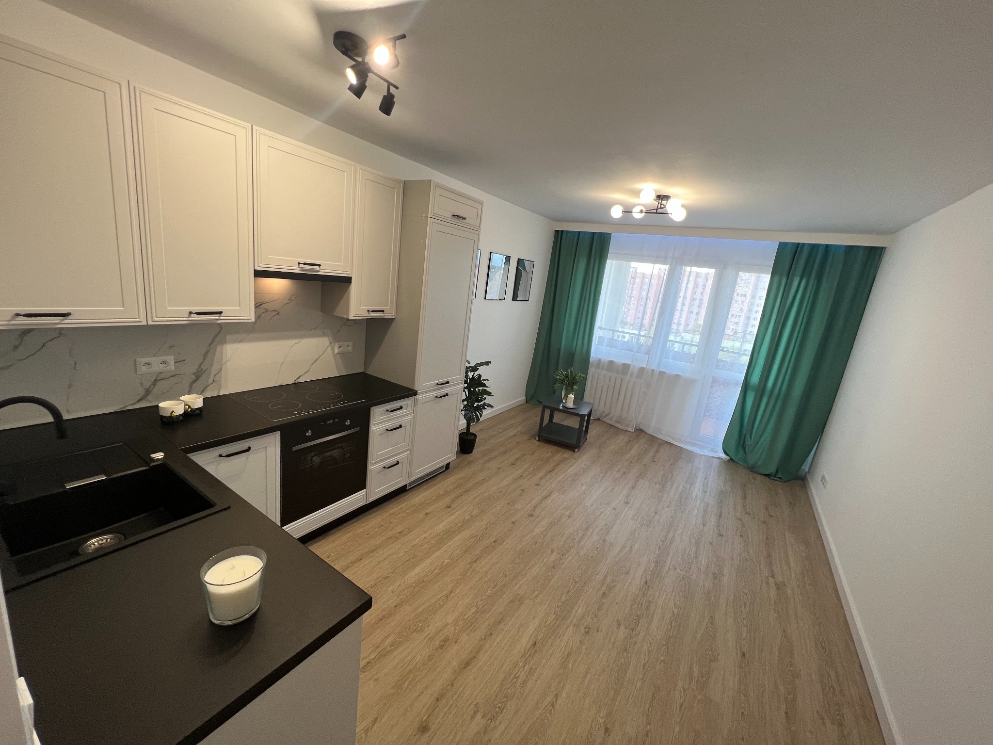 Jasna kuchnia z czarnym blatem, białymi szafkami i drewnianą podłogą, widok na okno z zielonymi zasłonami, nowoczesne oświetlenie sufitowe i dekoracje.