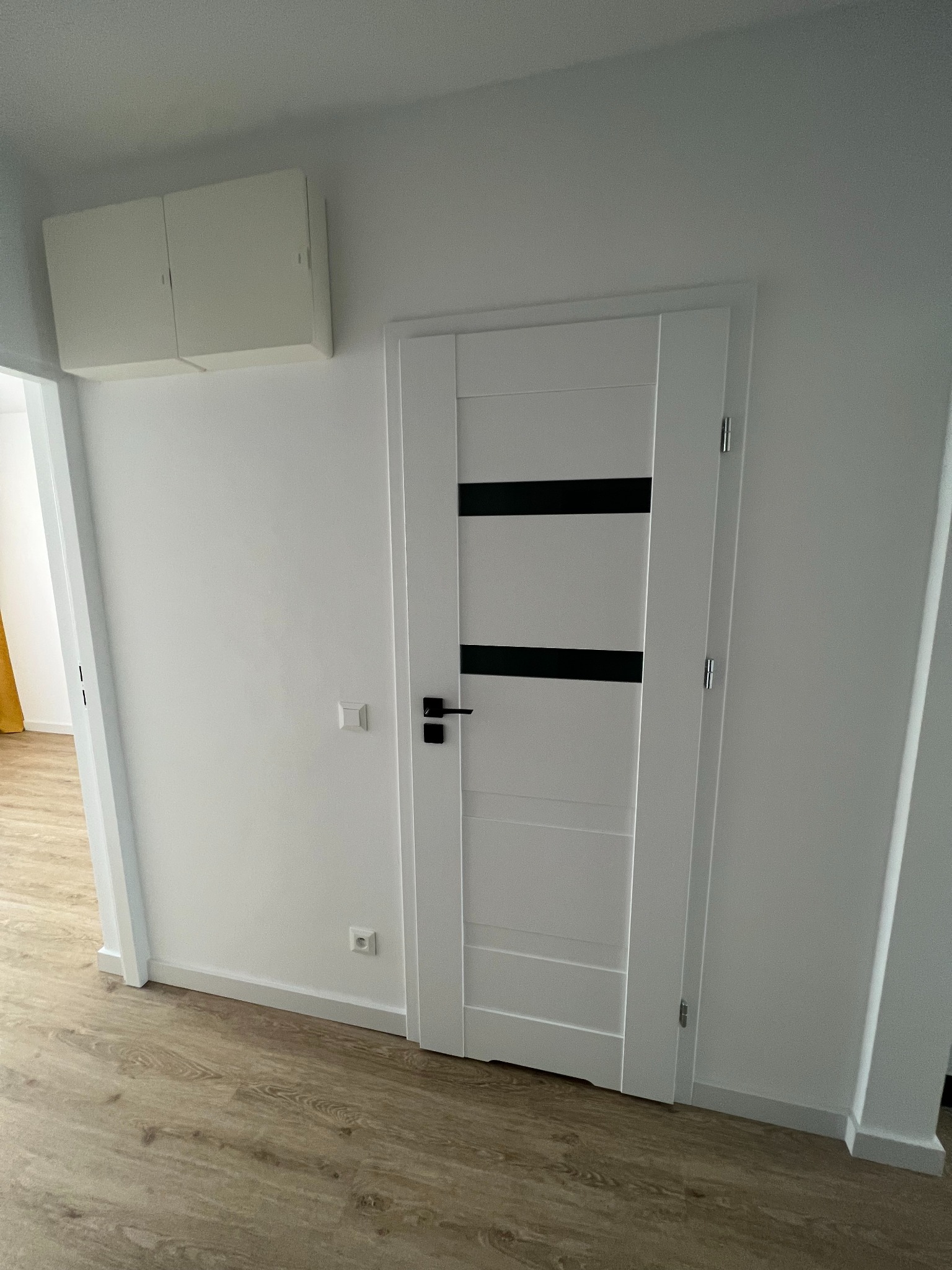 Nowoczesne, białe drzwi wewnętrzne z czarnymi, poziomymi akcentami, zamontowane w jasnym pomieszczeniu z panelami podłogowymi i szafką na ścianie.