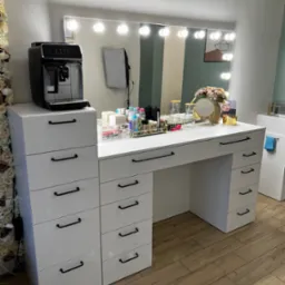 Biała toaletka z szufladami i lustrem otoczonym żarówkami, na blacie akcesoria do makijażu, ekspres do kawy, dekoracja kwiatowa i drewniana podłoga.