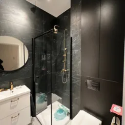 Nowoczesna łazienka z czarnymi marmurowymi płytkami, kabiną prysznicową z czarną ramą i złotą armaturą, białą szafką pod umywalkę z złotymi uchwytami i okrągłym lustrem.