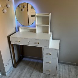 WOOD DESIGN Hubert Śmiglewski - Biała toaletka z trzema szufladami po prawej stronie i trzema szufladami pod blatem, okrągłe lustro z niebieskim podświetleniem LED, złote gałki szuflad, ustawiona na szarym panelu podłogowym.