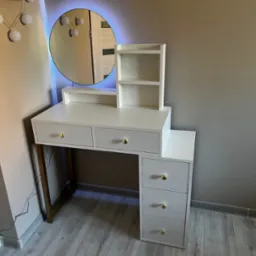 Biała toaletka z trzema szufladami po prawej stronie i trzema szufladami pod blatem, okrągłe lustro z niebieskim podświetleniem LED, złote gałki szuflad, ustawiona na szarym panelu podłogowym.