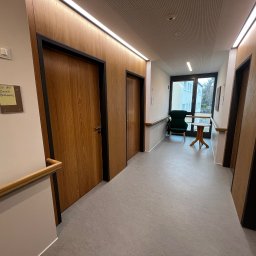 WOOD DESIGN Hubert Śmiglewski - Korytarz z drewnianymi drzwiami z czarnymi ościeżnicami, drewnianą poręczą i widokiem na zewnątrz przez okno na końcu korytarza.