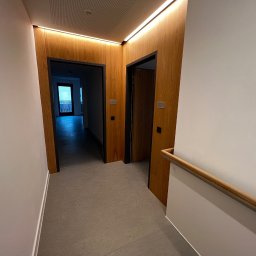 WOOD DESIGN Hubert Śmiglewski - Korytarz z dwoma drewnianymi drzwiami z czarnymi ościeżnicami, podświetlony listwą LED, z widoczną balustradą schodową i fragmentem balkonu w oddali.