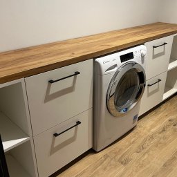 WOOD DESIGN Hubert Śmiglewski - Białe szafki z drewnianym blatem, wkomponowane w przestrzeń z pralką Candy, czarne uchwyty, podłoga imitująca drewno.