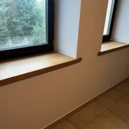 Dwa drewniane parapety okienne o jasnym odcieniu drewna, zamontowane w oknach z czarnymi ramami na tle jasnych ścian i płytek podłogowych.